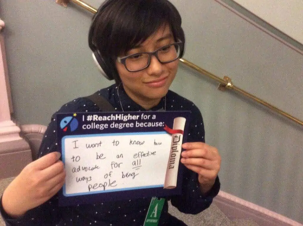 [Elly holds a sign reading "I #ReachHigher for a college degree because: I want to know how to be an effective advocate for all ways of being people"]