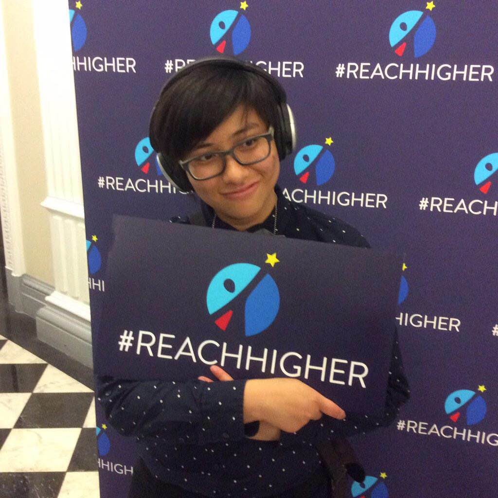 [A person named Elly Wong smiles and holds up a sign with the logo for the #ReachHigher campaign.]