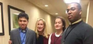 A young Asian man, a young white woman, and a young Black man are standing next to a white woman in her forties. They are in a hotel hallway.