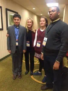 A young Asian man, a young white woman, and a young Black man are standing next to a white woman in her forties. They are in a hotel hallway.