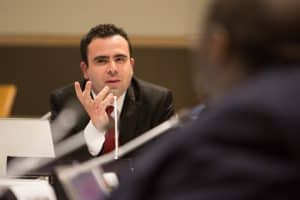 Photo of a man in a suit speaking at a table with a microphone