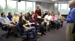 Photo of a group of people seated in chairs with one standing to ask a question to a man at the front of the room