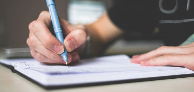 A person writing in a notebook with a blue ballpoint pen.