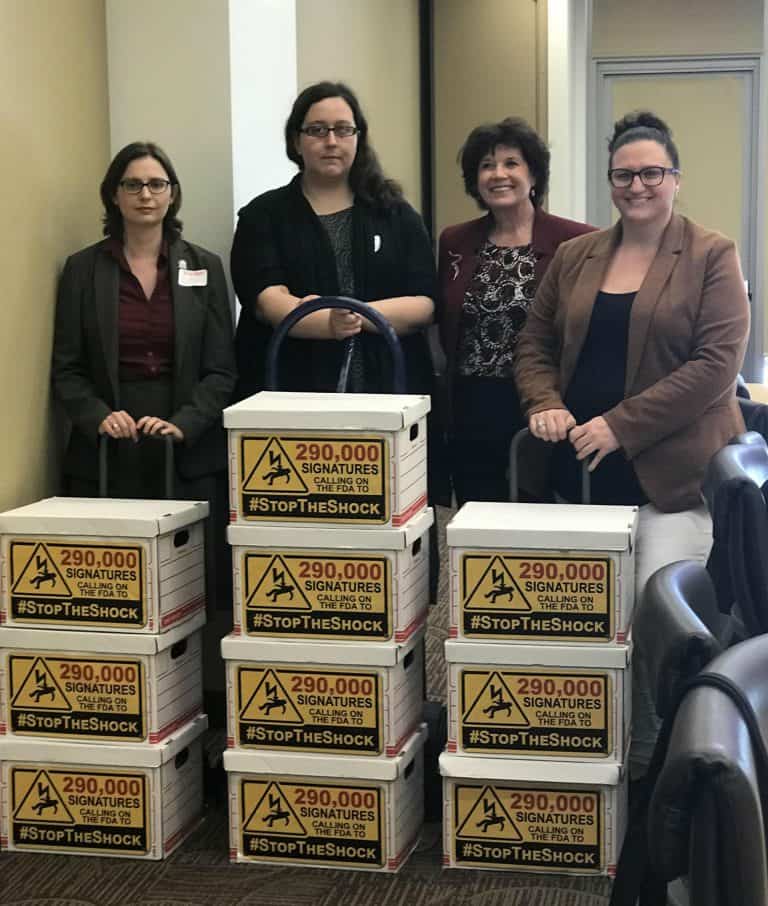 Sam Crane, Julia Bascom, Nancy Weiss, and Nicole Jorwic stand behind 3 stacks of white boxes on dollies. The signs on the boxes say "290,000 Signatures calling on the FDA to #StopTheShock".