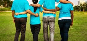 Photo of four people from behind. They are wearing blue shirts, have their arms around each other's waists, and are walking across grass