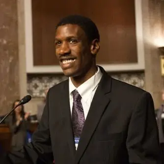 A smiling Black man dressed in a suit and tie stands at a podium delivering a speech with several people in the background