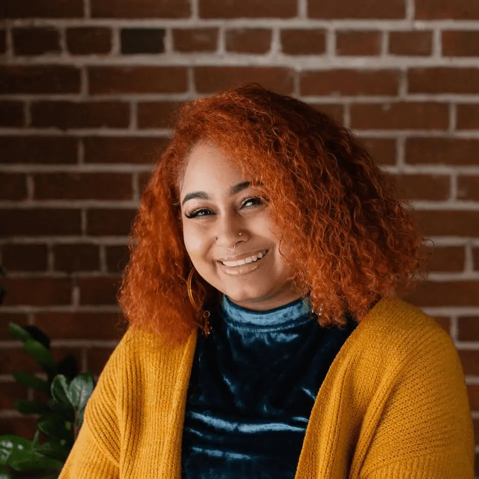 A smiling Black person in front of plants and a brick wall. She is tilting her head and looking at the camera. She has orange curly hair, two nose piercings, and large hoop earrings. Photo by Trista Marie photography.