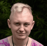 A young white person stands against a dark background, smiling at the camera. They have dyed blond hair that is shaved on the sides.