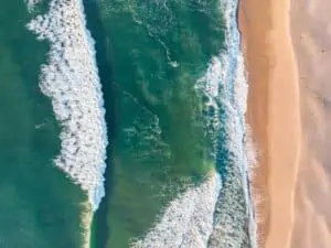 A birds-eye view of teal waves crashing onto a sandy beach
