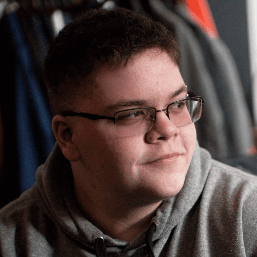 A photo of Gavin Grimm looking off to the right with a reflective expression on his face. Gavin has pale skin and short dark hair. He is wearing a light grey hoodie and half rim wire glasses. The background behind him is a mixture of greys and dark blues. He is lit starkly, so deep shadows are cast on the left side of his face.