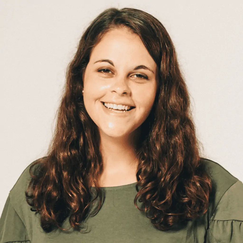 A photo of Jordyn Zimmerman, looking out at the audience and smiling with her head tilted slightly to the right. Jordyn has light olive skin and long slightly wavy medium brown hair that frames the sides of her face. She is wearing an olive green shirt and is in front of a pale background.