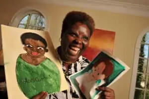Lois Curtis, a Black woman with short hair, smiles joyfully while holding up two paintings she has made. One is a self-portrait of Lois in a green shirt, labeled “Lois Curtis.” The other painting shows a woman with short brown hair and large green earrings.
