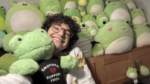 Jenna, a white person with short curly brown hair and glasses, holds a frog plush wearing an emotional support frog shirt. There are many frog plushes behind them.