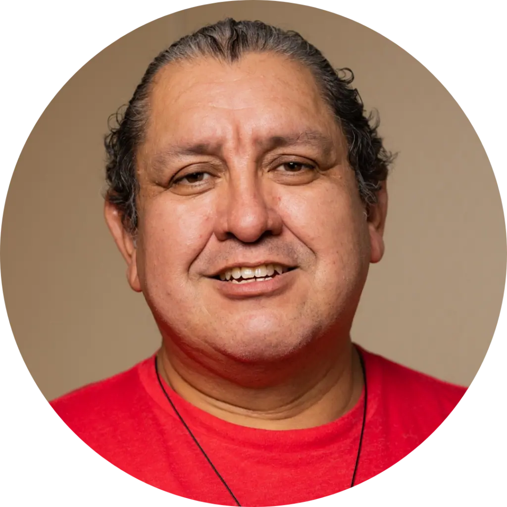 Héctor Manuel Ramírez, a Two-Spirit person with tan skin and short, dark curly hair pushed back.They wear a necklace and a red shirt with with the United Farm Workers union logo.