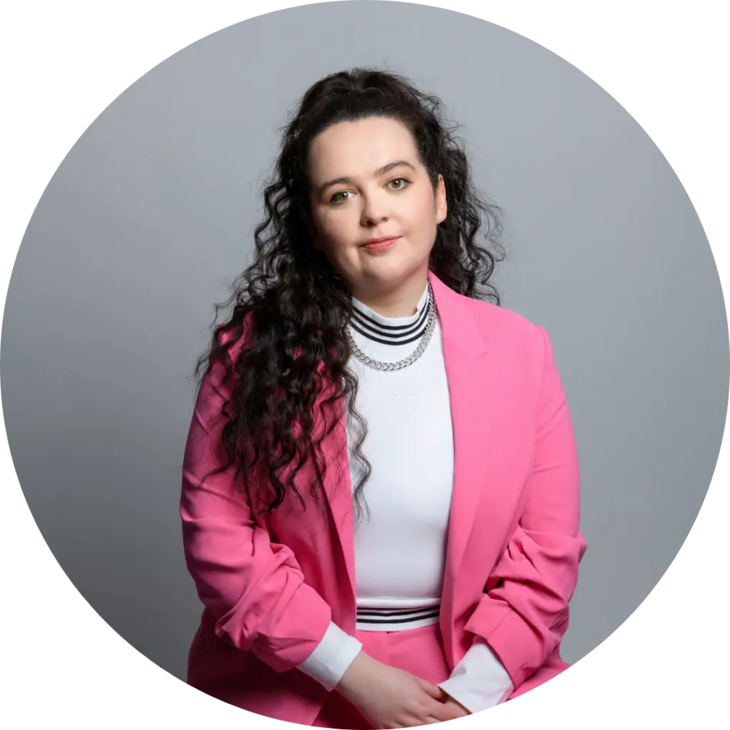 Ashley Storrie, a white woman with long curly brown hair. She wears a pink suit, white top, and green and white socks and shoes. She is sitting and holding her hands.